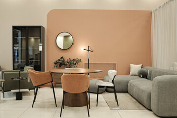 Modern furniture showroom displaying contemporary sofa, armchairs, round dining table and chairs arranged against peach accent wall with decorative mirror and indoor plant in retail setting
