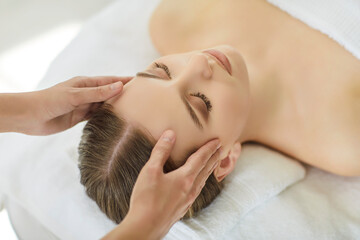 Young female lying on table with eyes closed during rejuvenating face massage, skilled therapist gently massages her head, close up photo. Self-care rituals, relaxation, tension relief, spa treatment 