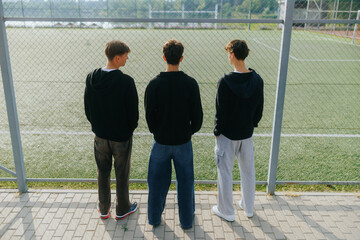 Three Teen Boys Standing by the Fence at a School Sports Field
