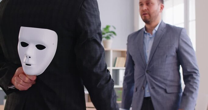 Untruthful fraudulent tricky immoral business man in suit with mask behind back pretends friendly at professional teamwork negotiation, makes false promise and fools company. Fraud, dishonesty concept