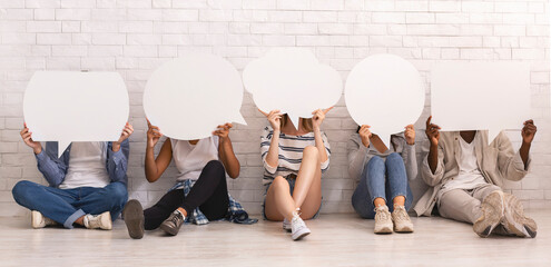 Group of young people sitting on floor over white brick wall, covering faces with speech bubbles, panorama