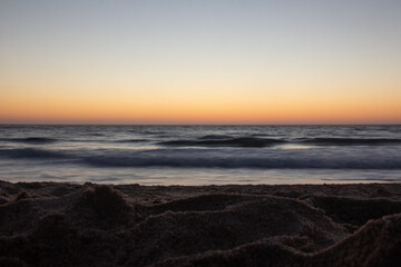 An artistic seascape captured with long exposure at sunset. The image is taken low to the ground, showing the sandy beach in the foreground and the blurred waves in motion.
