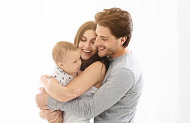 This image shows a happy family of three, a man, a woman, and their baby, standing close together in a white interior setting. The man and woman are smiling at the baby