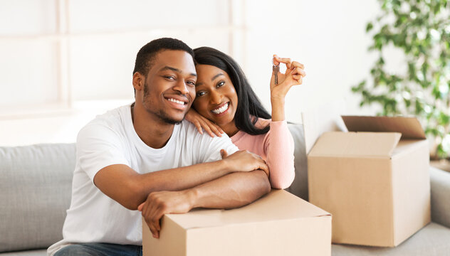 Happy black couple with house key and carton boxes looking at camera and smiling in their property, panorama. Young African American homeowners excited to move into new apartment - Powered by Adobe