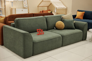 Green sofa standing on showroom floor with decorative pillows arranged on seat, red 50 percent...