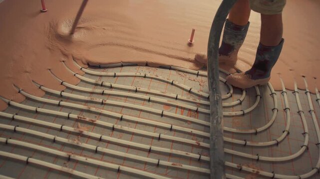 Worker pouring self-leveling concrete mix over underfloor heating pipes