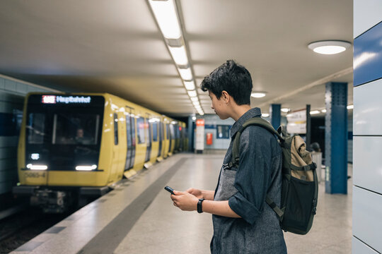 Teenager student girl with backpack waiting for the subway train and using a smartphone on the platform - Powered by Adobe