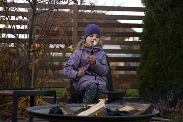 An eight-year-old girl roasts marshmallows on an autumn day near a country house.  Family leisure with kids at autumn.