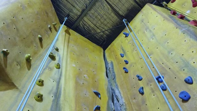 A static upward-facing shot of an indoor rock climbing wall featuring colorful holds, climbing ropes, and a belay pulley system. 