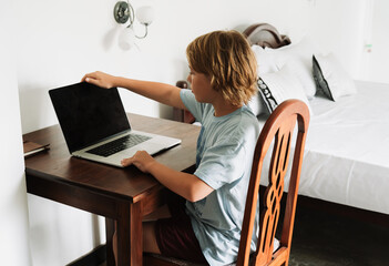 Teenage boy studying with a laptop in a bright tropical room in Sri Lanka. Digital nomad lifestyle, online learning, and early IT education. Cozy workspace with natural light and wooden furniture