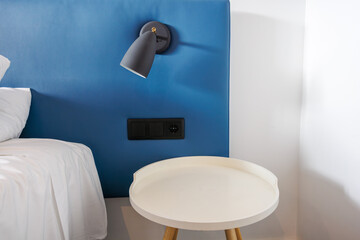 Close-up of modern grey metal lamp, bedside table in contemporary bedroom. White room interior with bed with blue headboard and white bedding.