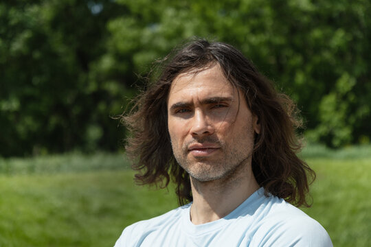 Portrait of handsome caucasian brunette man with long curly hair and dissatisfied emotion and feeling unwell at park and green nature setting. Summer overheating in sun day. Slow living. - Powered by Adobe
