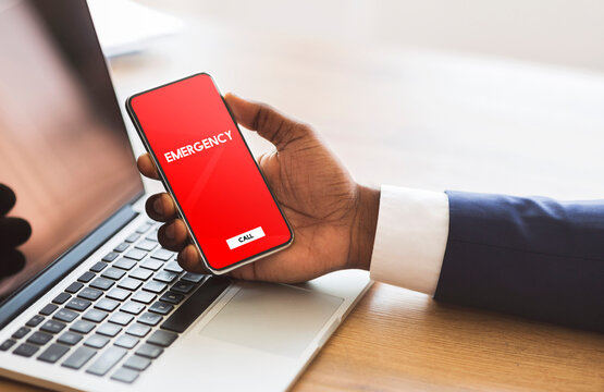 Safety Concept. Male hand holding smart phone with emergency number on red screen over office background and laptop