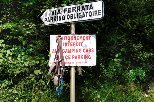 white sign with VIA-FERRATA PARKING OBLIGATOIRE and STATIONNEMENT INTERDIT AUX CAMPING CARS PARKING in French, Via Ferrata access information, sign indicating mandatory parking for route