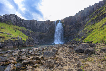 Majestic waterfall flowing down rugged basalt cliffs into clear river in Iceland. Scenic view showcasing Iceland's untouched natural beauty, lush greenery, dramatic geology and powerful cascades.