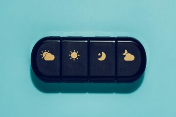 Black daily pill organizer with gold sun, cloud, moon icons, centered on vibrant blue. Symbolizes health, wellness, medication management, and daily routines.