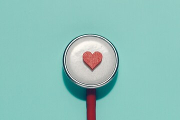 Small red heart rests centrally on a silver stethoscope diaphragm against a blue green background. Symbolizes healthcare, cardiology, heart health, medical care, and compassion in medicine.