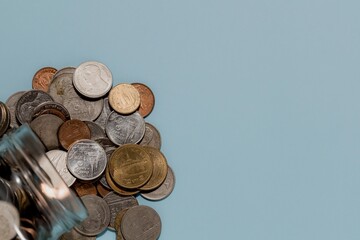 Thai Baht coins spill from a glass jar onto light blue with copy space. Symbolizes saving, financial management, currency, and money.