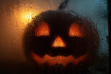 Close-up of foggy jack-o'-lantern face glowing faintly through wet glass window, droplets and reflection visible