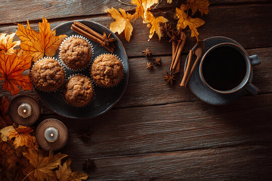 Interior flat lay of a dark wooden kitchen table with freshly baked pumpkin muffins, cinnamon sticks, star anise, fall leaves, and a ceramic cup of coffee, candles glowing nearby
