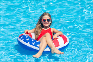 Happy smiling little girl 7-8 years old in red swimsuit and sunglasses relaxing in outdoor swimming pool on lifebuoy decorated with American flag print. Vacation in USA concept. 4th of July pool party