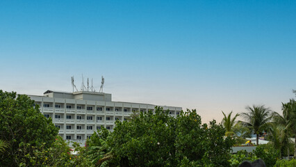 Fototapeta premium Tropical urban landscape with a mid-rise building, telecommunication antennas on the roof, and lush green trees under a clear blue sky at sunrise.