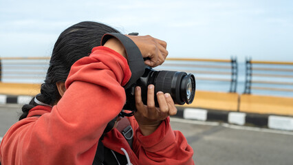 Woman photographer in red hoodie taking a photo with DSLR camera near oceanfront road, creative hobby concept with lifestyle and travel vibes.