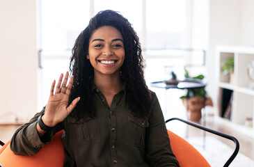Hello. Portrait Of Cheerful Young African American Woman Waving Hand Smiling To Camera Sitting On Pouf Chair At Home. Hi Greeting Gesture Concept. Webcam Shot, Video Call Screen, Remote Communication