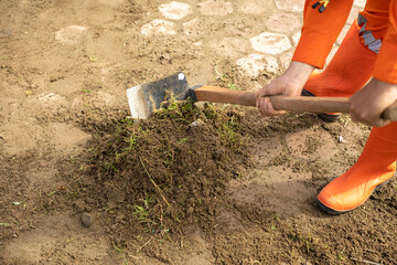 Worker in bright orange uniform using a hoe to dig and loosen soil on the ground during outdoor manual labor or cleanup activity.