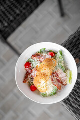 Fresh Caesar salad with bacon, cherry tomatoes, croutons, and Parmesan in a white bowl on a wicker chair with tiled background, top view, close-up.