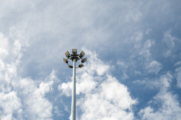 A tall stadium floodlight pole stands against a bright blue sky with scattered clouds. Minimalist background for energy, public lighting, and infrastructure concepts.
