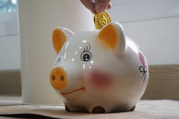 Hand places gold coin into cheerful ceramic piggy bank, symbolizing savings and financial growth. piggy bank is white with orange accents and painted heart, sitting on wooden surface