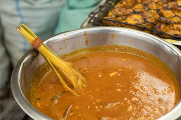Traditional yellow curry sauce in a large metal bowl with lemongrass tied as a natural brush, used for basting grilled food at a street food stall.