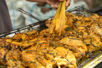 Yellow seasoned chicken being grilled in iron mesh, basted with lemongrass brush, highlighting traditional Indonesian barbecue technique and aromatic turmeric spice.