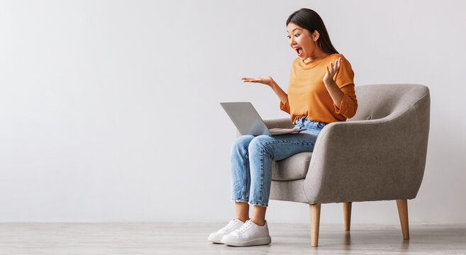 Emotional young Asian lady sitting in armchair with laptop, shouting OMG, celebrating success against white wall, banner with copy space. Excited woman enjoying big sale or online win - Powered by Adobe