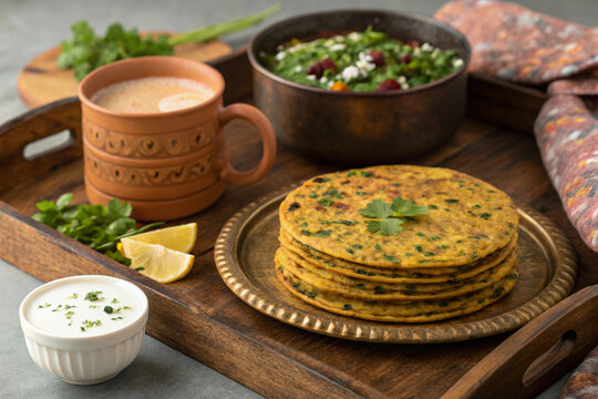 A delicious Indian breakfast spread featuring freshly made methi thepla stacked on a brass plate, served with a side of yogurt garnished with herbs, a steaming cup of chai in an earthen-style mug, and
