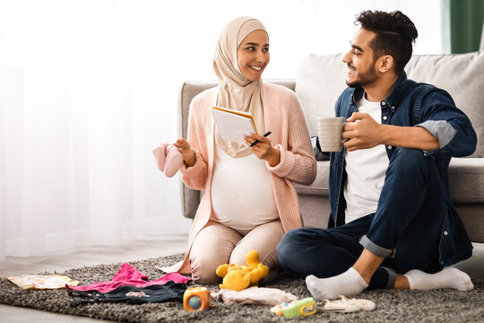 Happy pregnant muslim couple getting ready for childbirth, making checklist of necessities while sitting together on floor in living room with baby clothes around, islamic spouses relaxing at home - Powered by Adobe