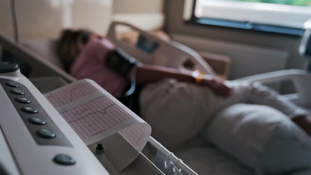 Close-up of a fetal monitor in a doctor's office, displaying fetal heart rate and uterine contraction data during a prenatal check-up with a blurred view of a pregnant woman lying in a hospital bed