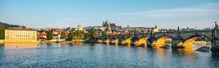 Breathtaking panorama showcases Prague Castle rising above the Vltava River, with the iconic Charles Bridge spanning the calm waters, reflecting the morning light in a serene cityscape.