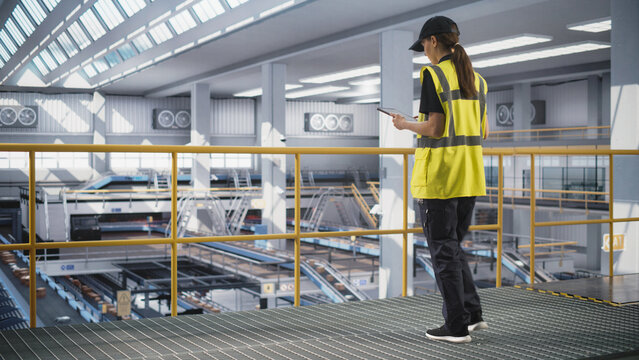 Young Female Standing on a Metal Platform, Using a Tablet Computer at a Post Office Logistics Center. Specialist Working at a Sorting Facility, Configuring the Automation Process