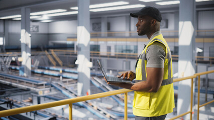 Black Employee Working at a Modern Logistics Center for an International Online Shopping Business. African Man Using a Laptop Computer to Optimize the Orders Before Deliveries