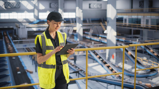 Female Working in a Modern Advanced Logistics Center with Automated Belt Conveyors for Sorting Packages. Young Woman in High Visibility Vest Using Laptop Computer in an Industrial Facility