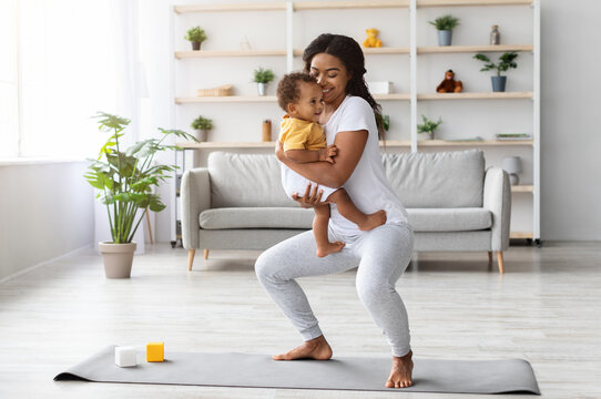 Postpartum Weight Loss. Happy Black Lady Making Squats With Baby On Hands While Training At Home, Smiling African American Woman Exercising In Living Room And Bonding With Infant Child, Copy Space - Powered by Adobe
