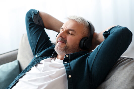 Joyful senior man reclining on couch with closed eyes, listening to music, using powerful wireless modern headphones, closeup, copy space. Modern technologies and entertainment concept - Powered by Adobe