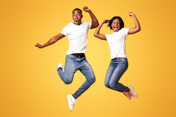 Happy black man and woman celebrating success, full length photo, copy space. Cheerful african american young couple clenching fists and screaming, jumping up over orange studio background