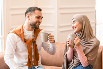 Happy Muslim Arab Couple Drinking Coffee Sitting On Sofa Smiling Each Other At Home. Cheerful Spouses Flirting Enjoying Conversation, Bonding Spending Time Together On Weekend