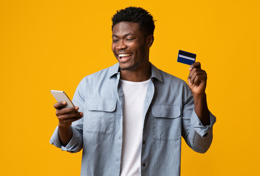 Cheerful african american young man holding brand new cellphone and credit card, trading or gambling online, recommending banking or shopping mobile app, yellow studio background, mockup, copy space