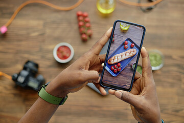 Black man holding smartphone capturing photo of grilled bread and cherry tomatoes on plate with food photography equipment and ingredients visible on table