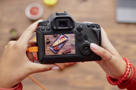 Young woman holding digital camera capturing overhead shot of baguette sandwich and cherry tomatoes on wooden table, hands visible adjusting camera settings during food photography session