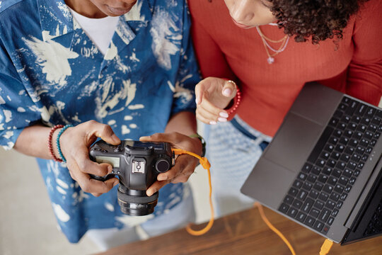 Young adult Black woman and young adult Black man reviewing digital camera images together while holding laptop, collaborating on food photography project, hands and torsos visible - Powered by Adobe